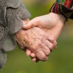 Two people are holding hands. One hand is older with visible veins running along the back of the hand.