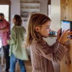 Home 14 A little girl looking closely at a digital screen placed on the wall inside a kitchen with her family in the background.