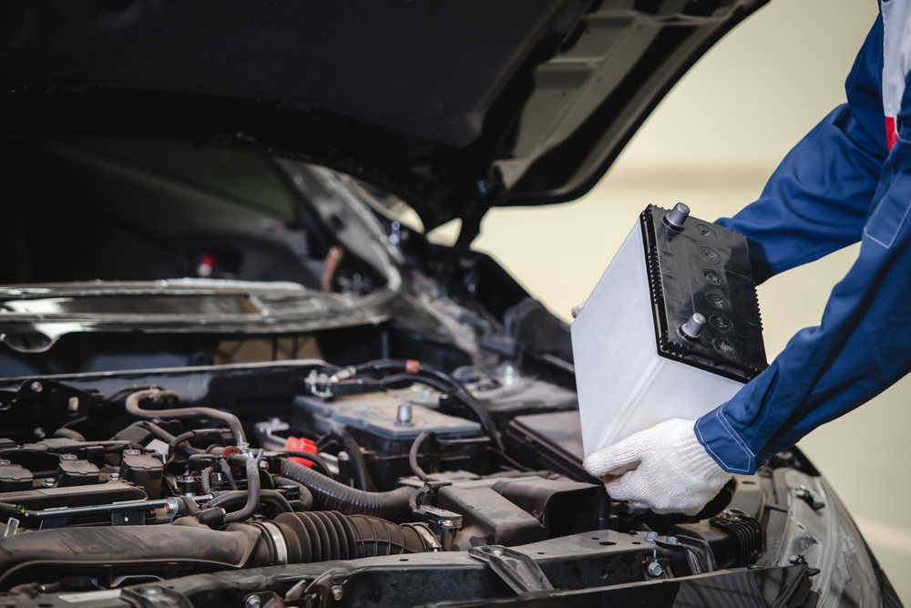 A mechanic wearing white gloves changes the battery of a car with its hood open, showing its components.