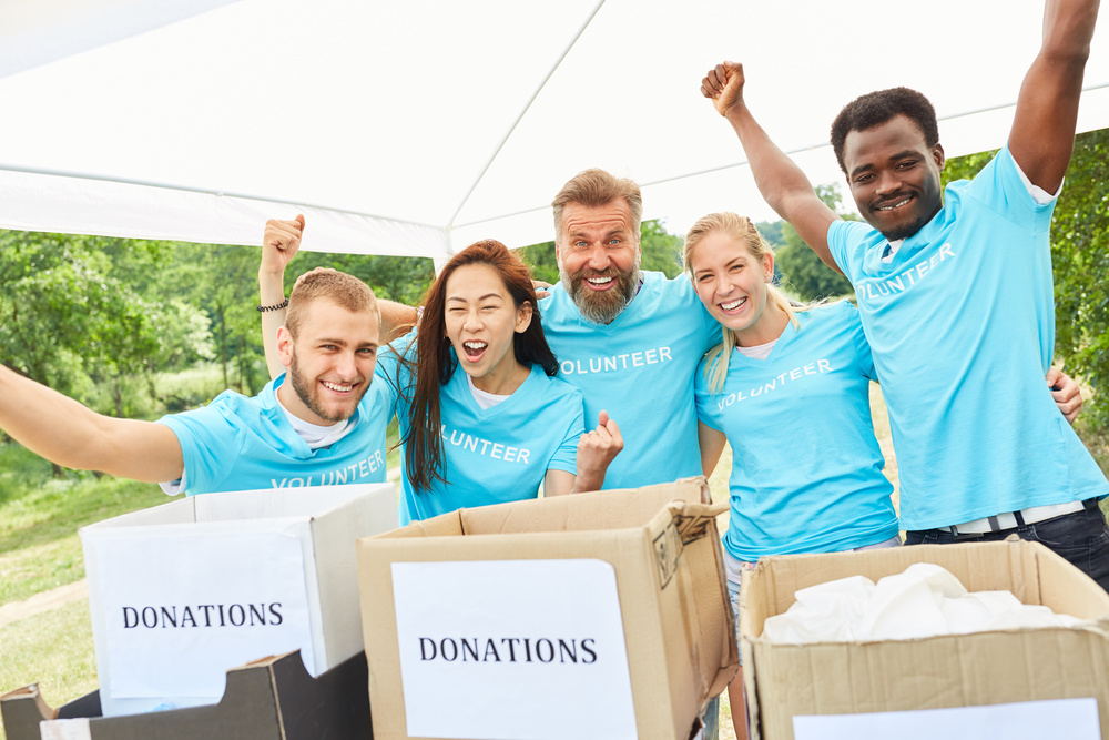 Home 11 Five people wearing blue volunteer shirts standing behind donation boxes, cheering enthusiastically.