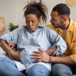 A pregnant woman sits on a couch, holding her belly, doing some deep breathing exercises. A man is behind her.