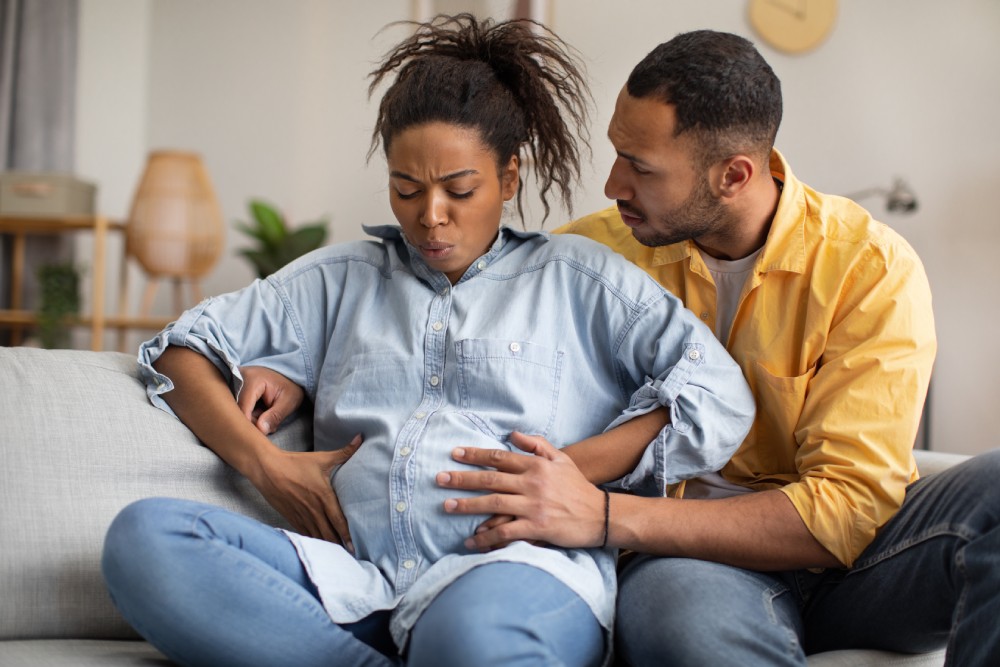A pregnant woman sits on a couch, holding her belly, doing some deep breathing exercises. A man is behind her.