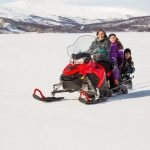 A woman and two children riding a red snowmobile on a snowy landscape, with mountains and trees in the background.