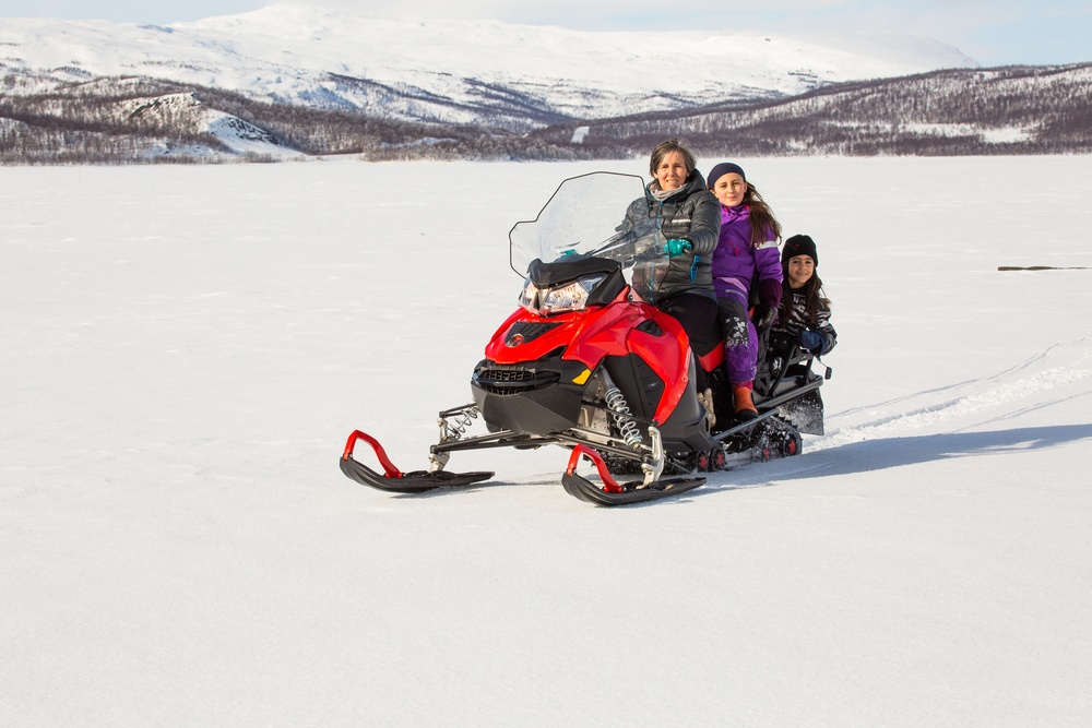 A woman and two children riding a red snowmobile on a snowy landscape, with mountains and trees in the background.