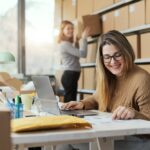 One woman is sitting in a chair looking at a piece of paper while a second woman is putting a box on a shelf.