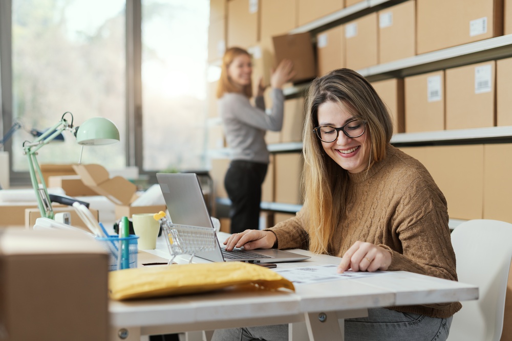 One woman is sitting in a chair looking at a piece of paper while a second woman is putting a box on a shelf.