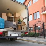 A truck with the back doors open, filled with furniture, boxes, suitcases, and plants, parked in front of a townhome.