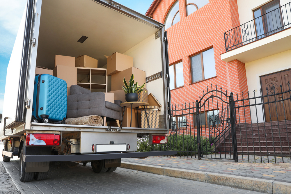 A truck with the back doors open, filled with furniture, boxes, suitcases, and plants, parked in front of a townhome.