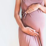 A woman wearing a simple pink dress standing against a white background, holding her pregnant belly.