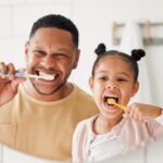 A young girl and an older man standing side-by-side in front of a bathroom mirror, smiling as they brush their teeth together.