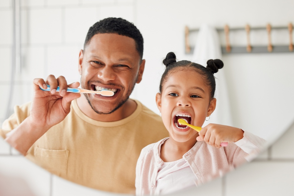 A young girl and an older man standing side-by-side in front of a bathroom mirror, smiling as they brush their teeth together.