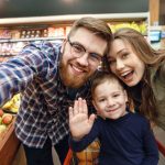 A Millennial man and woman smile as they pose with a child sitting in a shopping cart in a supermarket aisle.