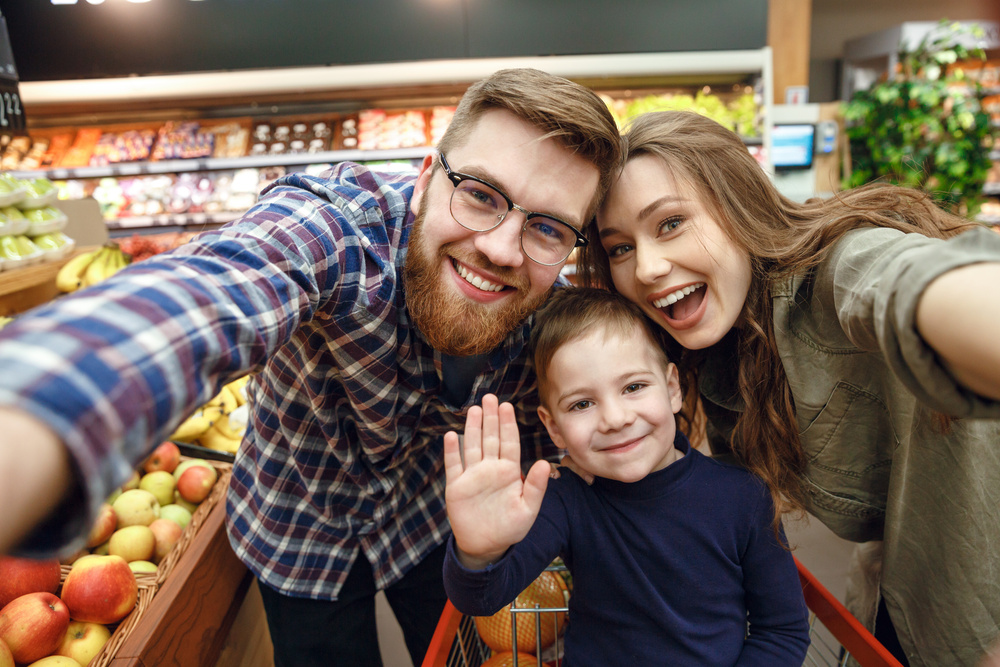 A Millennial man and woman smile as they pose with a child sitting in a shopping cart in a supermarket aisle.