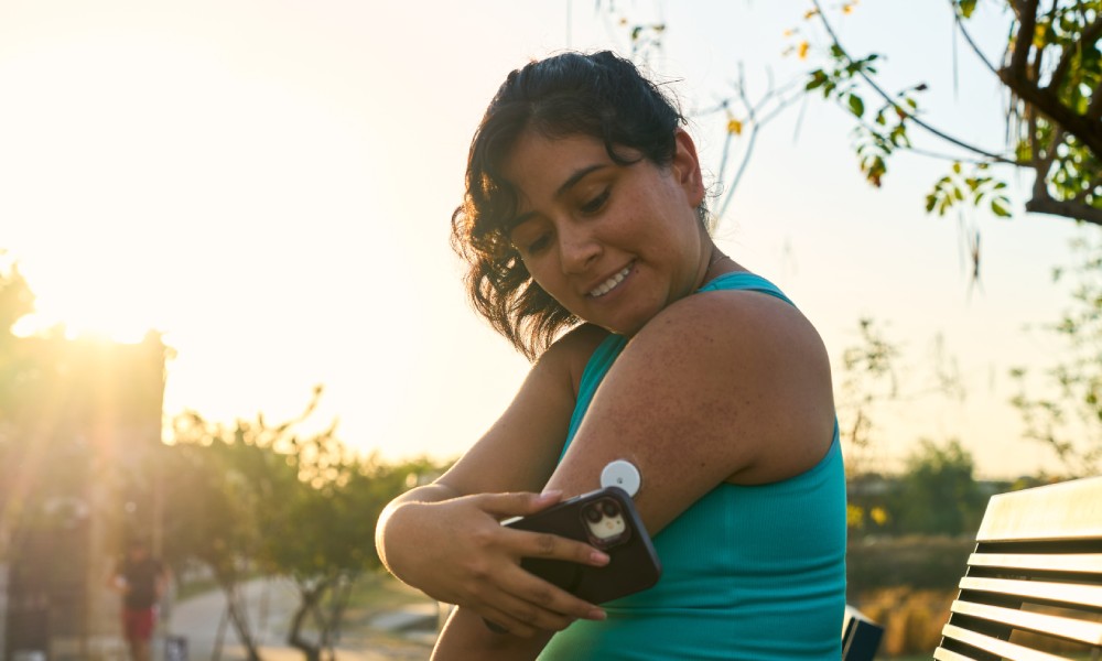 A woman sits on a park bench in workout clothes. She holds her phone up to a glucose monitor on her arm.