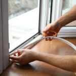 A close-up view shows a person wearing a white T-shirt installing a sealant strip across the bottom of a window.
