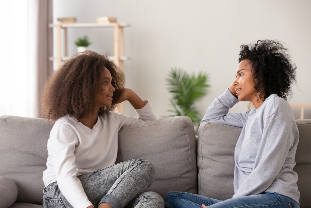 A young, female teen and her mom sit across each other on a gray couch while talking in the living room.