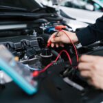 A technician using a small red tool connected to a blue device to repair a car battery in a brightly lit shop.