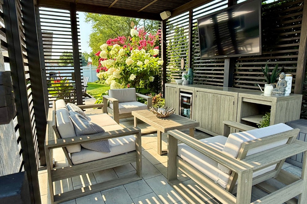 A shaded pavilion made of dark wood in a backyard space with outdoor seating arranged around a TV and console.