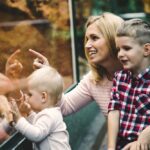 A smiling blonde mother with two children kneeling in front of a glass enclosure or display at a zoo.