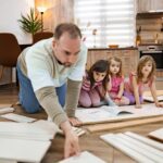 A man and three girls kneeling on the floor of a kitchen, with parts from an unbuilt piece of furniture in front of them.