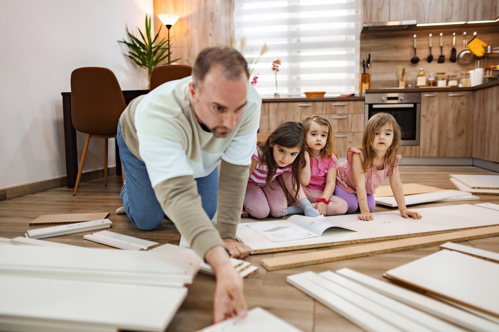 A man and three girls kneeling on the floor of a kitchen, with parts from an unbuilt piece of furniture in front of them.