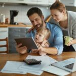 A father looks at a tablet device with his child as he sits and works in his kitchen. The mother stands behind him.