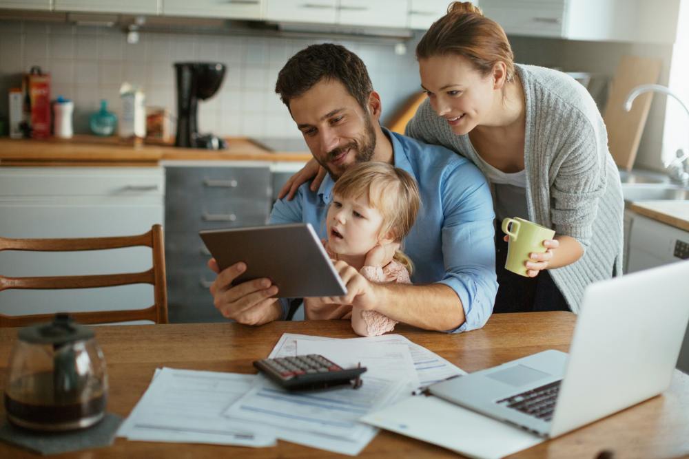 A father looks at a tablet device with his child as he sits and works in his kitchen. The mother stands behind him.