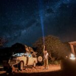 A man pointing a flashlight up to the starry sky, next to a pickup truck with a camping tent and a small fire pit.