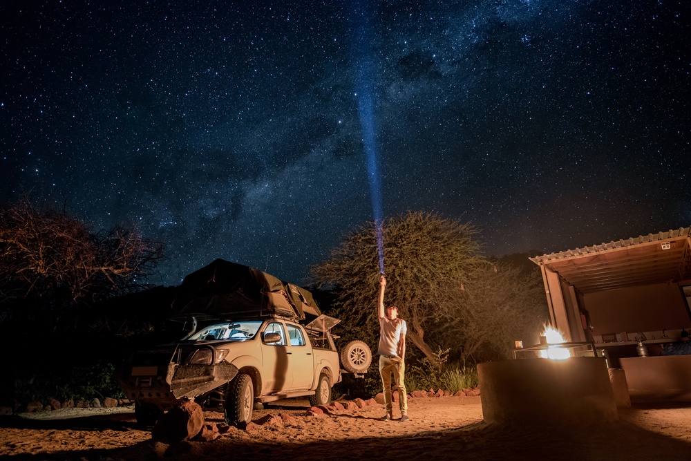 Home 24 A man pointing a flashlight up to the starry sky, next to a pickup truck with a camping tent and a small fire pit.