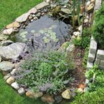 A backyard pond is bordered by large rocks, purple flowers, and lush greenery. A retaining wall is behind the pond.