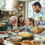 A family of five gathered around a dining table covered in food, from casserole to snacks like fruit and pretzels.