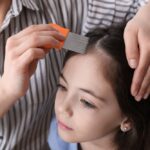 A woman using a fine-toothed nit comb on a young girl's hair as part of an anti-lice treatment while they both stand indoors.