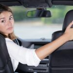 A woman in a car turning to look over her shoulder while reversing, one hand on the wheel and one on the passenger seat.