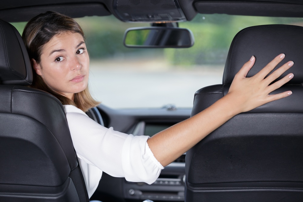 A woman in a car turning to look over her shoulder while reversing, one hand on the wheel and one on the passenger seat.